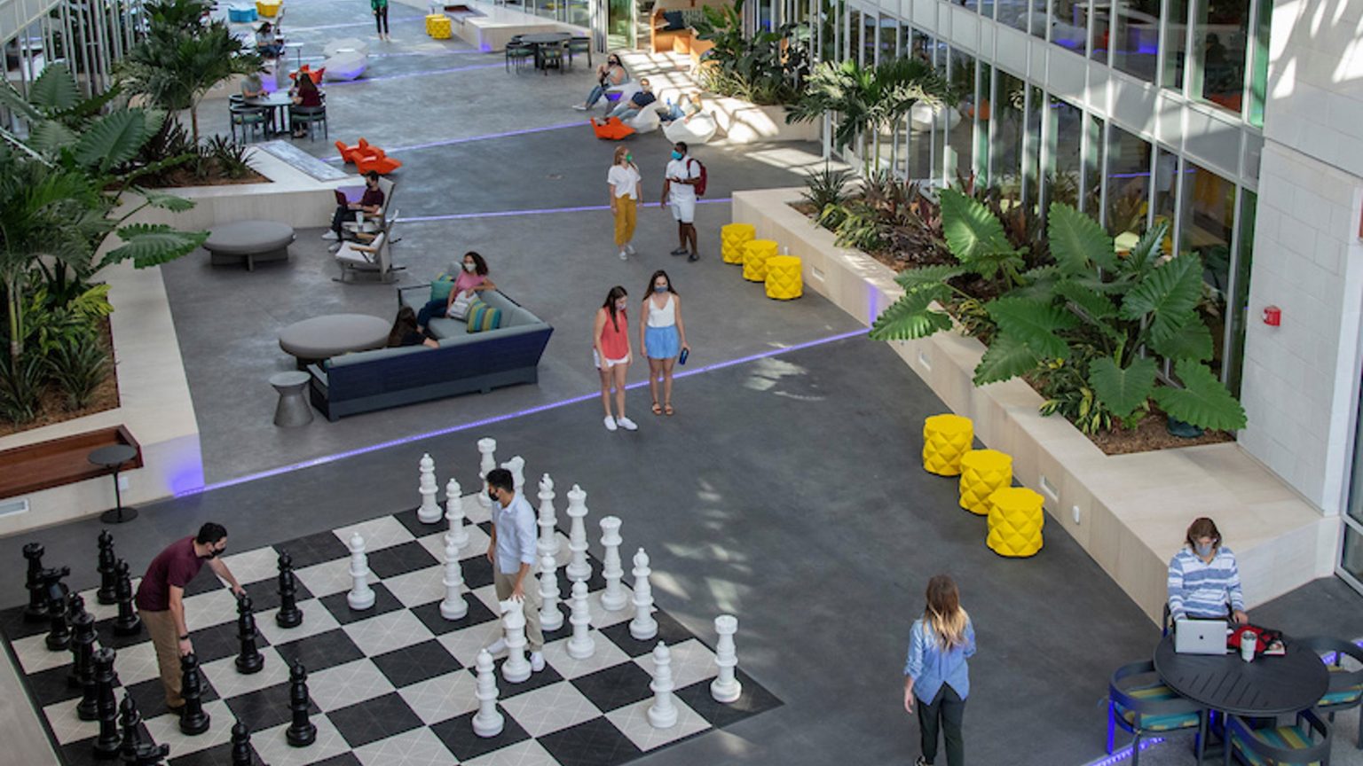 Overhead view of pedestrians moving through a mixed‑use complex featuring an outdoor chessboard, benches and colourful design elements.