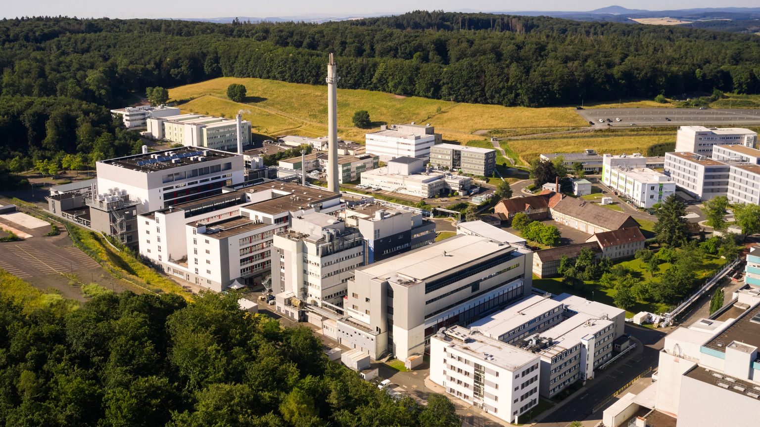 Drone perspective over a modern research and production facility nestled within dense woodland on the outskirts of a city.